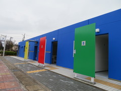 色分けされた屋外のトイレの写真。ウェルカムゲート横（無料エリア）に入口が黄色（車いすマーク）、赤色（女性）、青色（男性）のトイレがあります