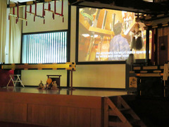 ステージ奥のスクリーン（字幕付）の写真。英語字幕付きの「からくり人形の説明映像」が流れています。